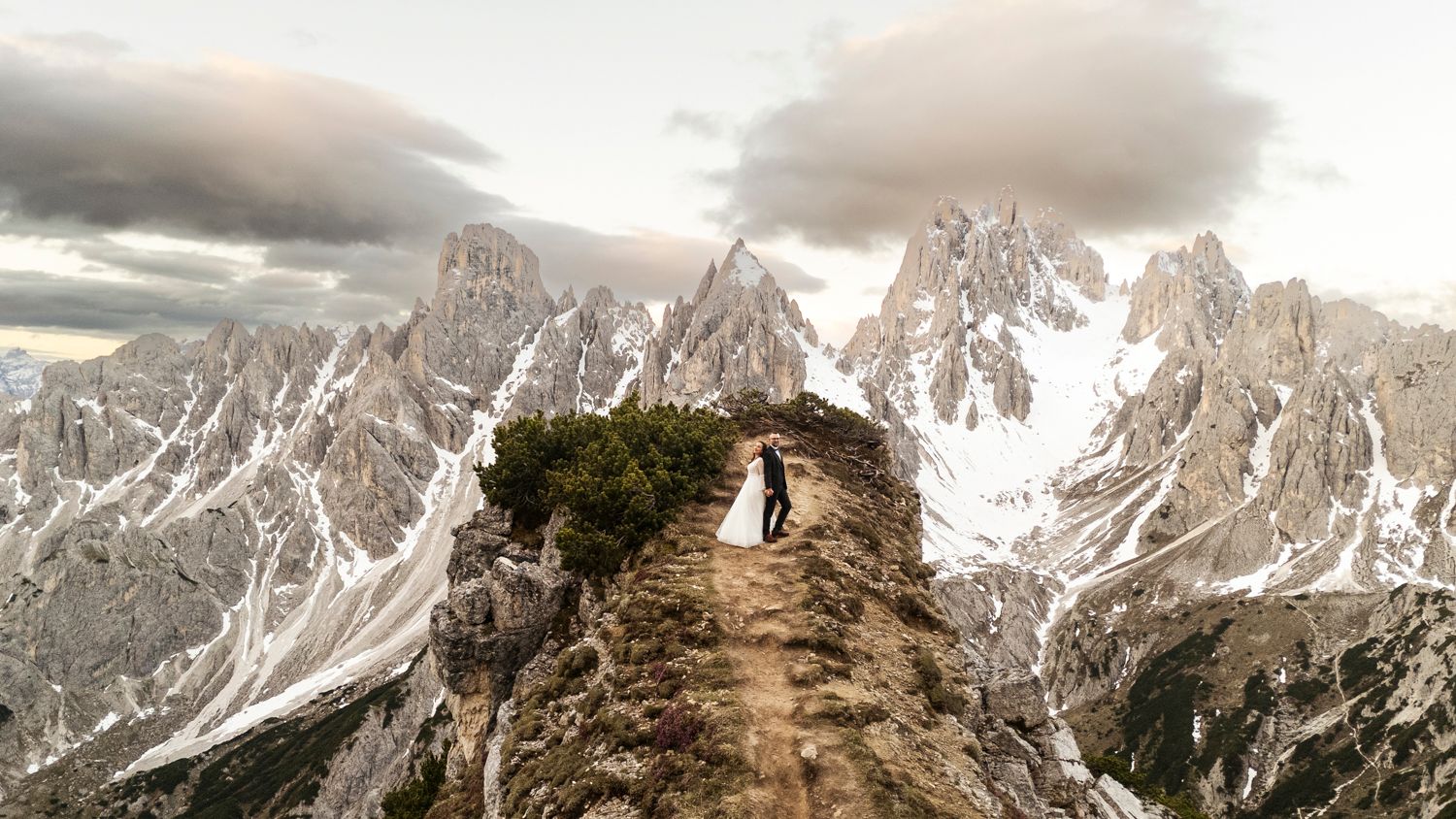 Silhouetted figures stand on narrow mountain ridge path with dramatic peaks behind.