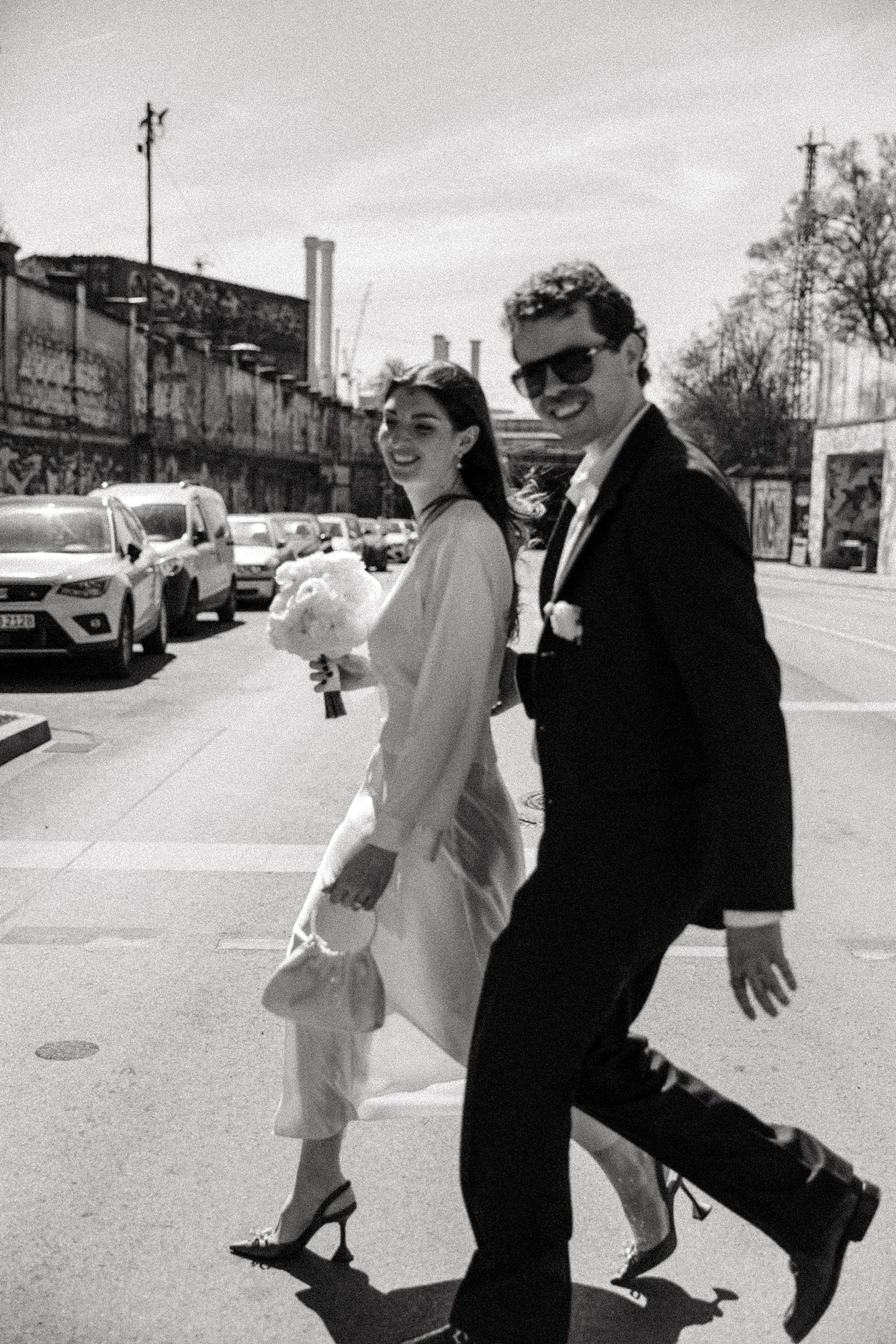 A stylish couple walking down a city street in wedding attire carrying a white bouquet.