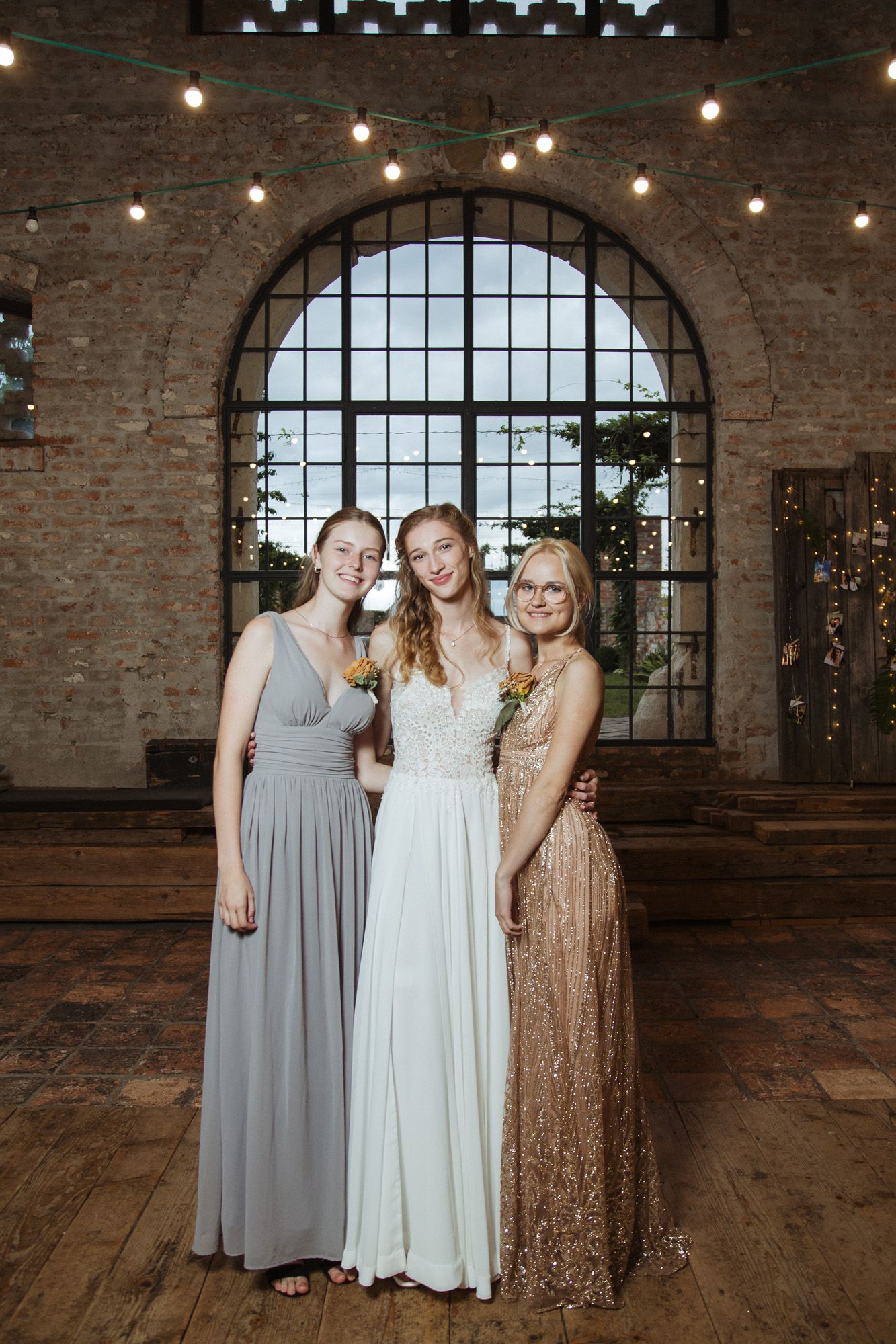 Three wedding attendees pose in elegant dresses inside a rustic venue with an arched window and string lights.