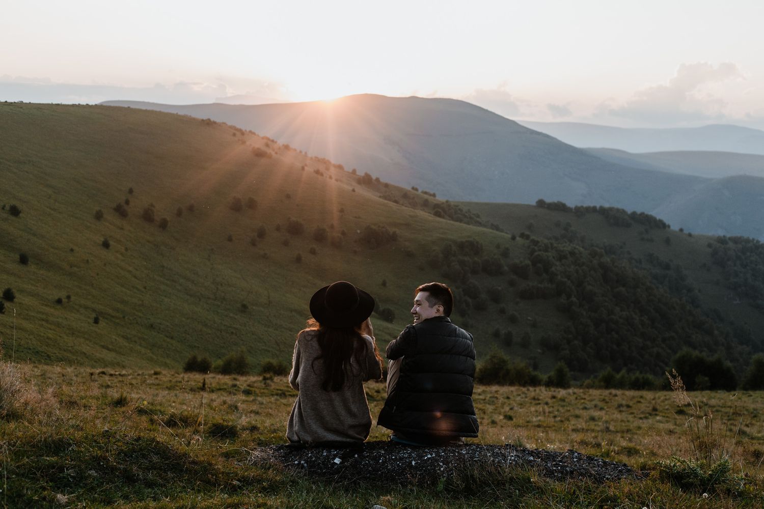 Silhouettes sitting on hillside watching golden sunset over mountain range.
