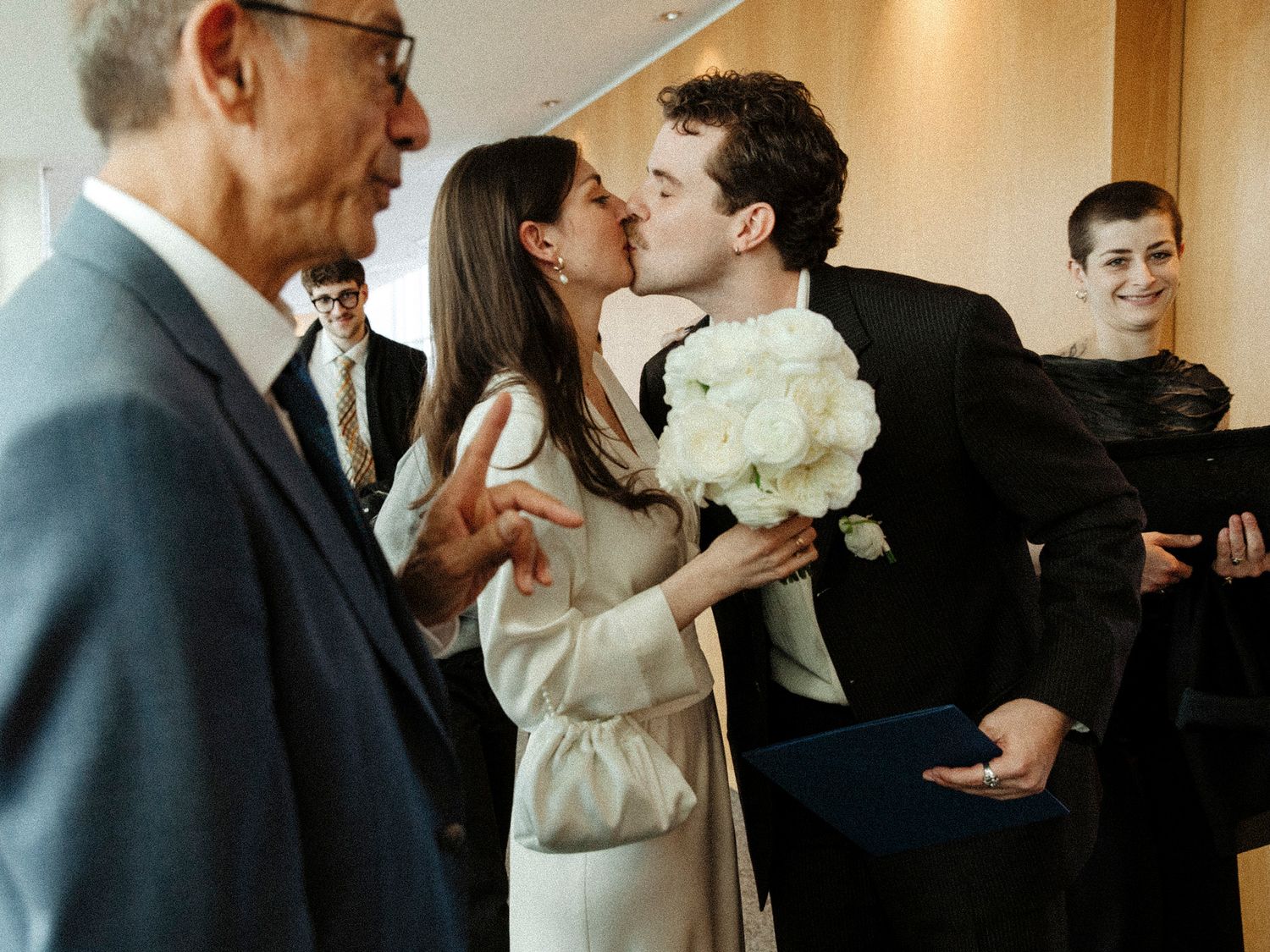A candid wedding moment with guests watching as the couple shares a kiss while holding white flowers.