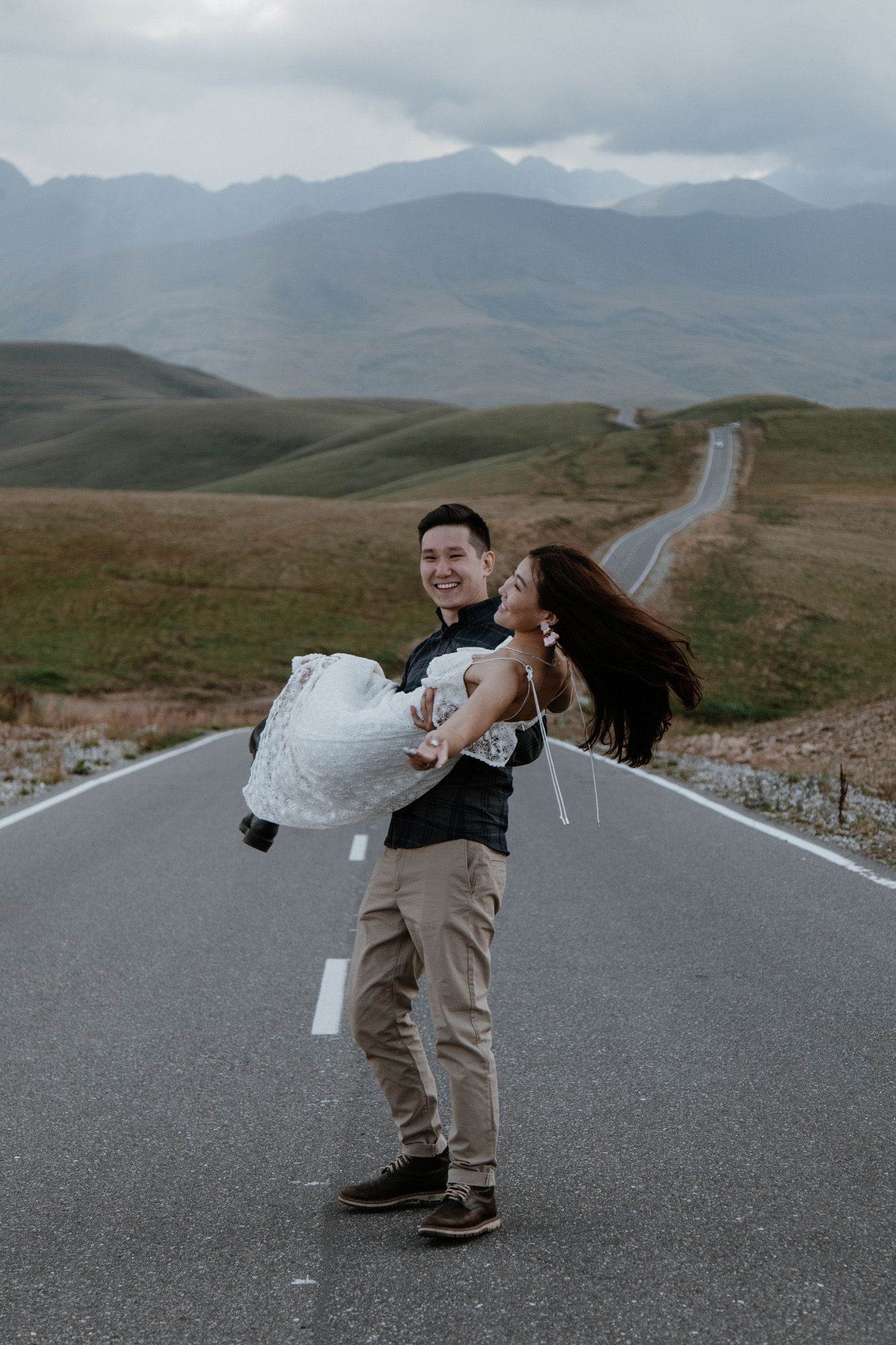 A couple shares a playful moment on a scenic mountain road with dramatic landscapes in the background.