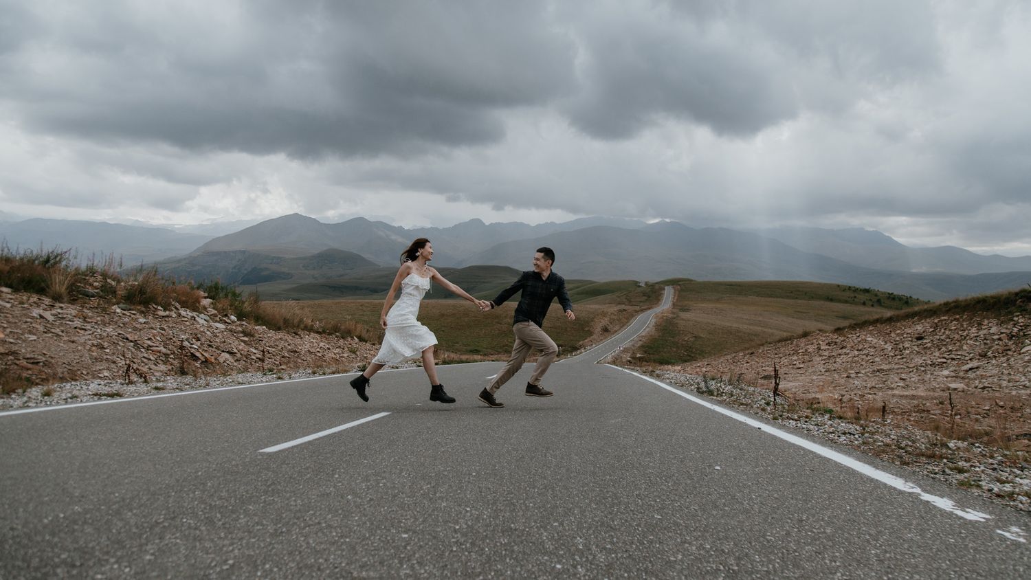 Two people dance playfully on an empty mountain road with dramatic landscape views.