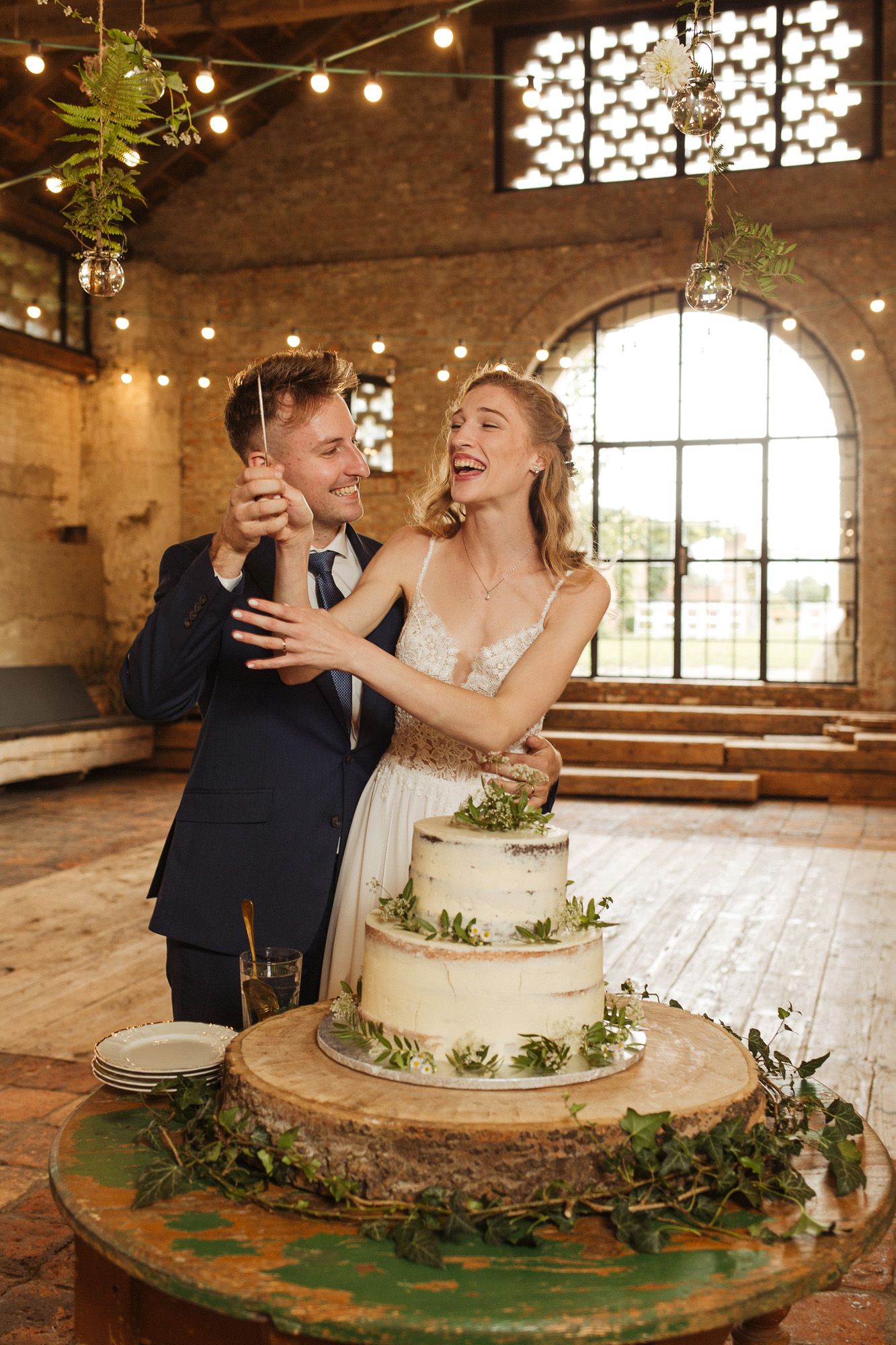 Wedding cake cutting moment in rustic venue with decorative window and string lights.