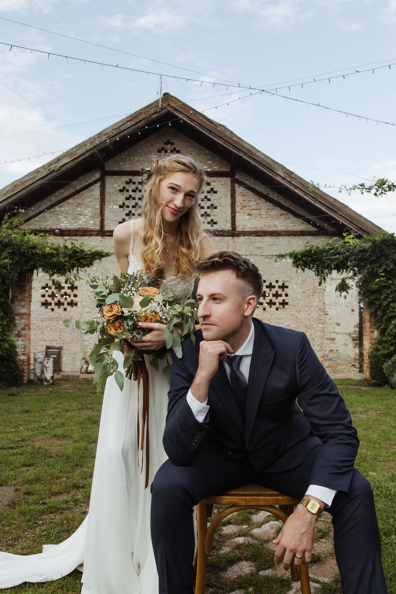 Wedding couple poses creatively with wooden chair against vintage brick building facade.