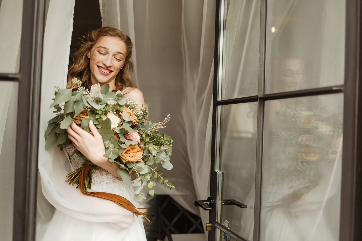 Candid bridal moment with lush bouquet of roses and eucalyptus by window.