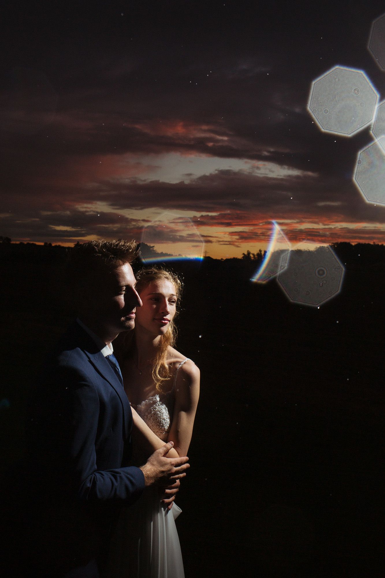 Artistic wedding portrait at sunset with dramatic lighting and lens flares.