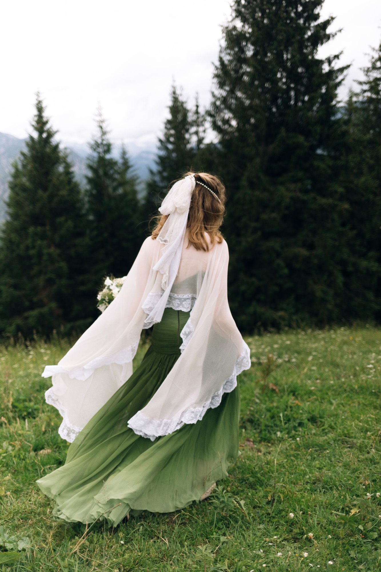 A bride in a flowing olive green and white wedding dress twirls on a hilltop meadow with evergreen trees in the background.