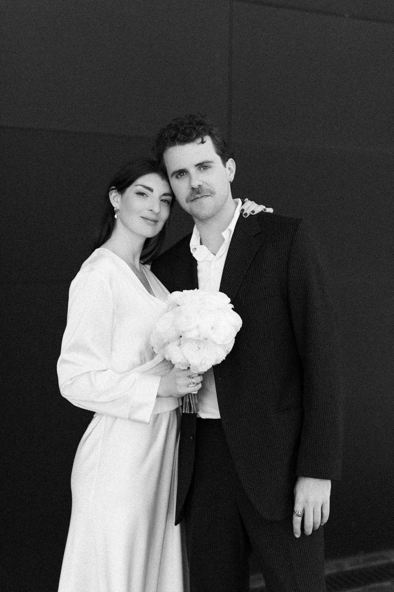 Black and white portrait of couple in formal attire with white bouquet against dark background.