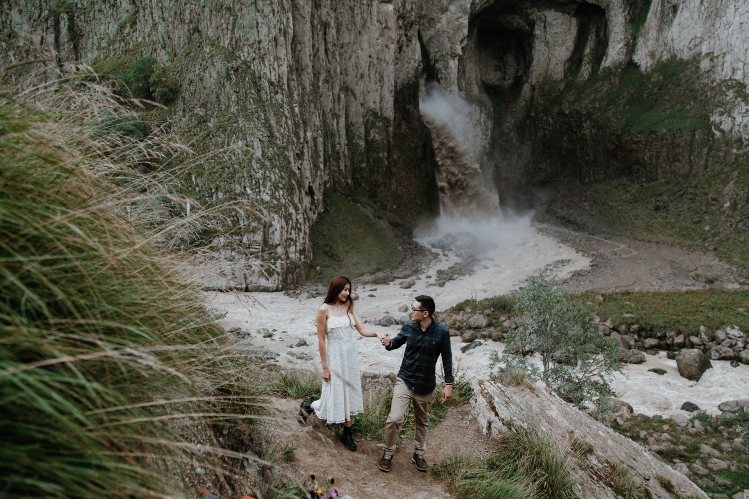 Couple explores dramatic waterfall canyon with towering limestone cliffs and flowing water in background.