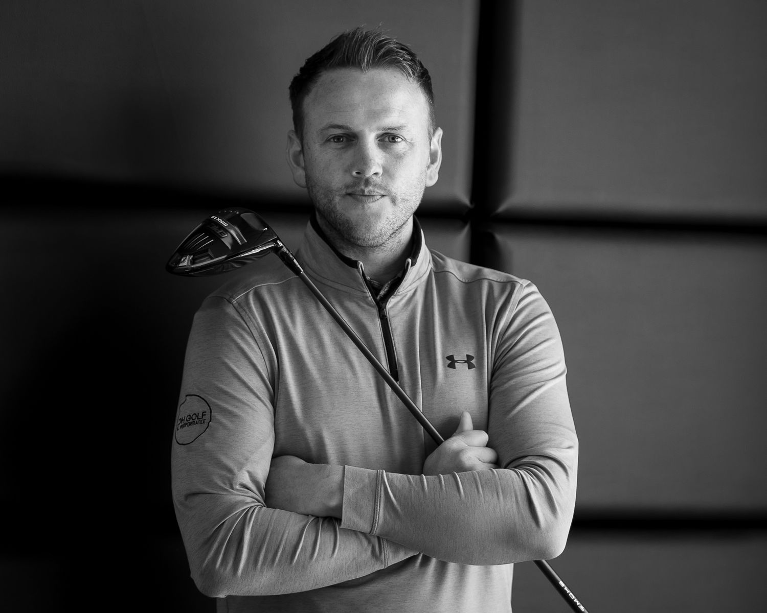 Athletic coach in Under Armour jacket poses with crossed arms in monochrome portrait.