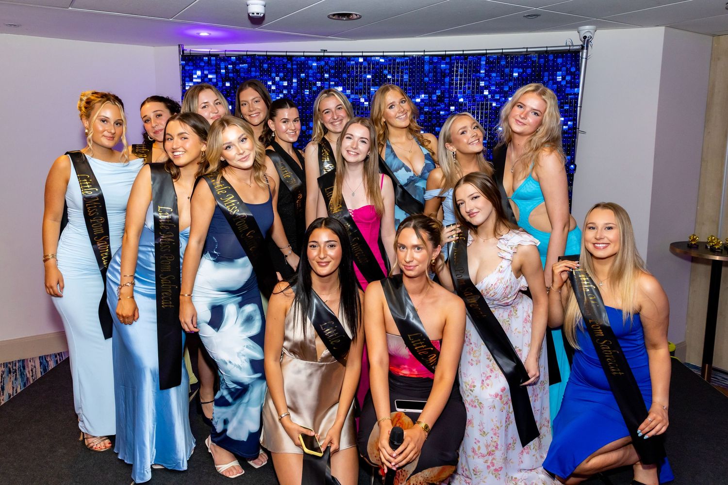 A group wearing sashes and formal attire poses together against a sparkly blue backdrop at an event.