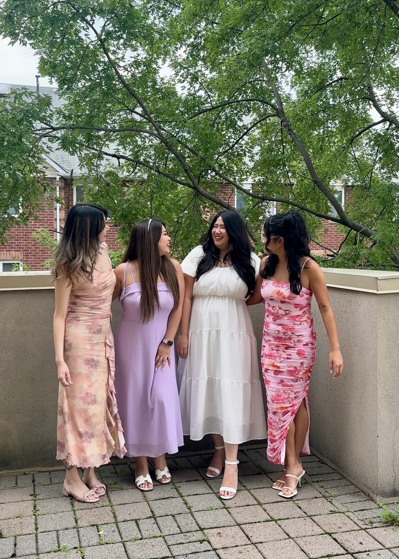 Four friends in colorful dresses pose together on a stone patio with green trees in the background.