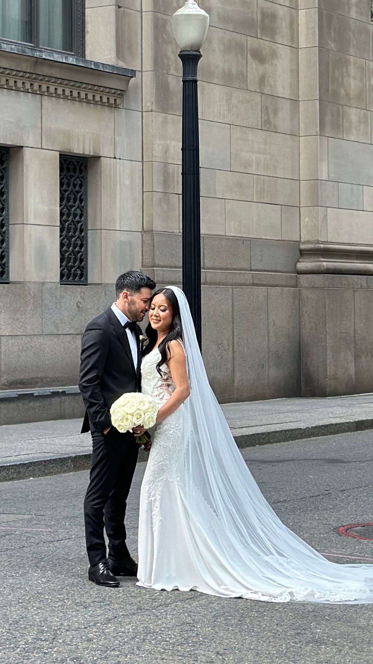 Classic wedding portrait with couple against stone building and lamppost in urban setting.