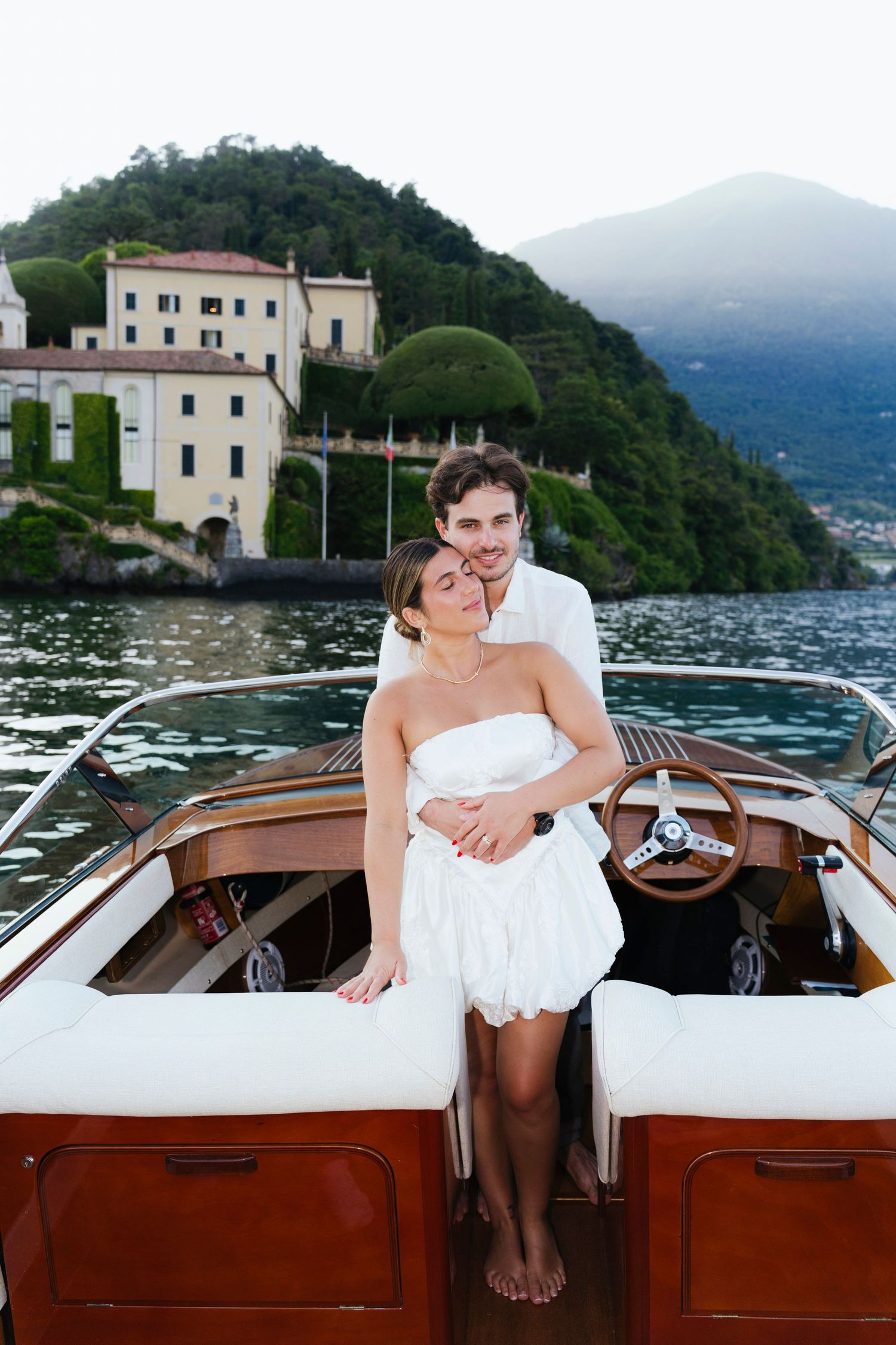 Luxury wooden boat cruising along Italian coastline with Villa Balbianello visible on Lake Como's mountainous shore.