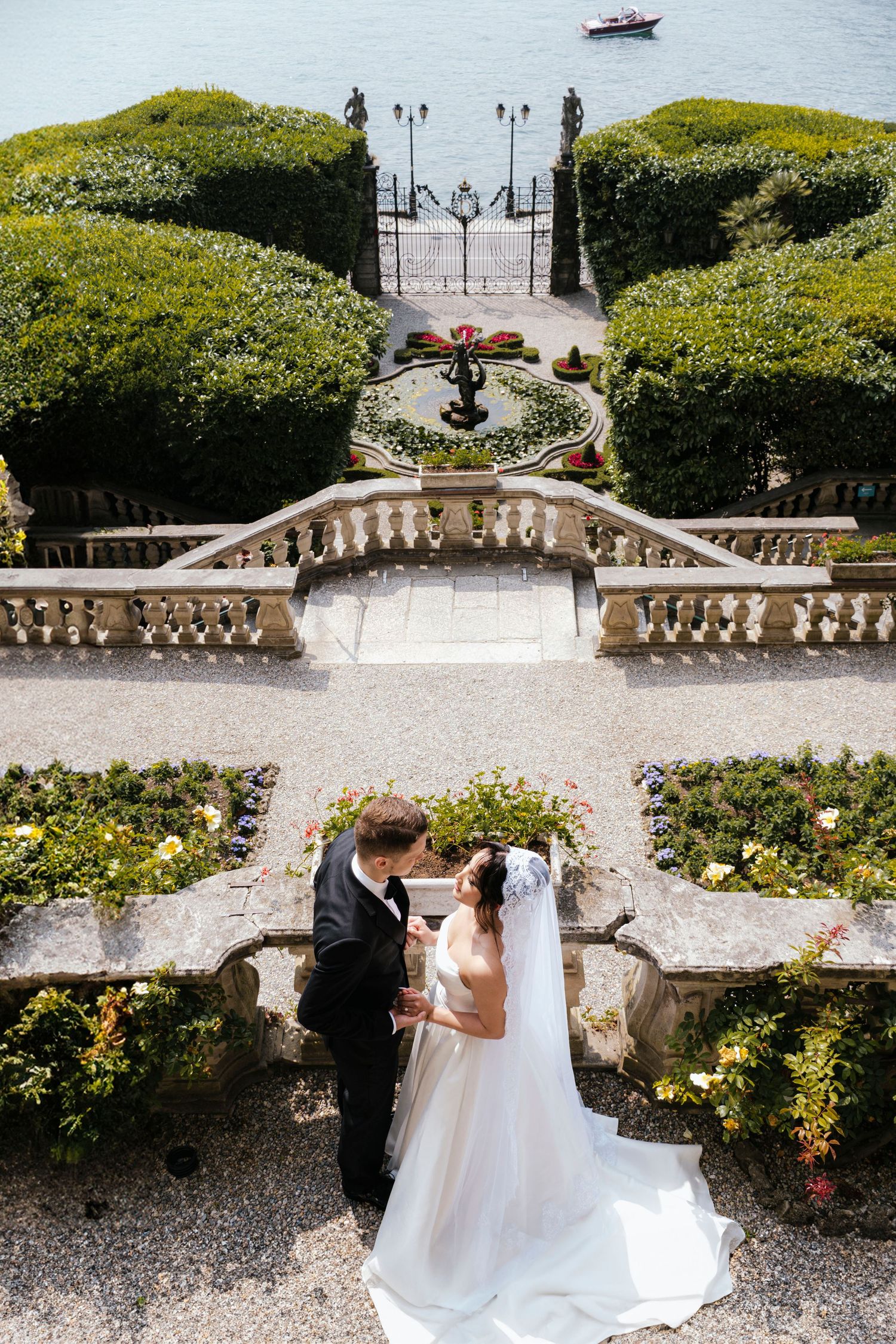 Couple stands together in elegant wedding attire on ornate stone terrace overlooking lakefront garden with boat in distance.
