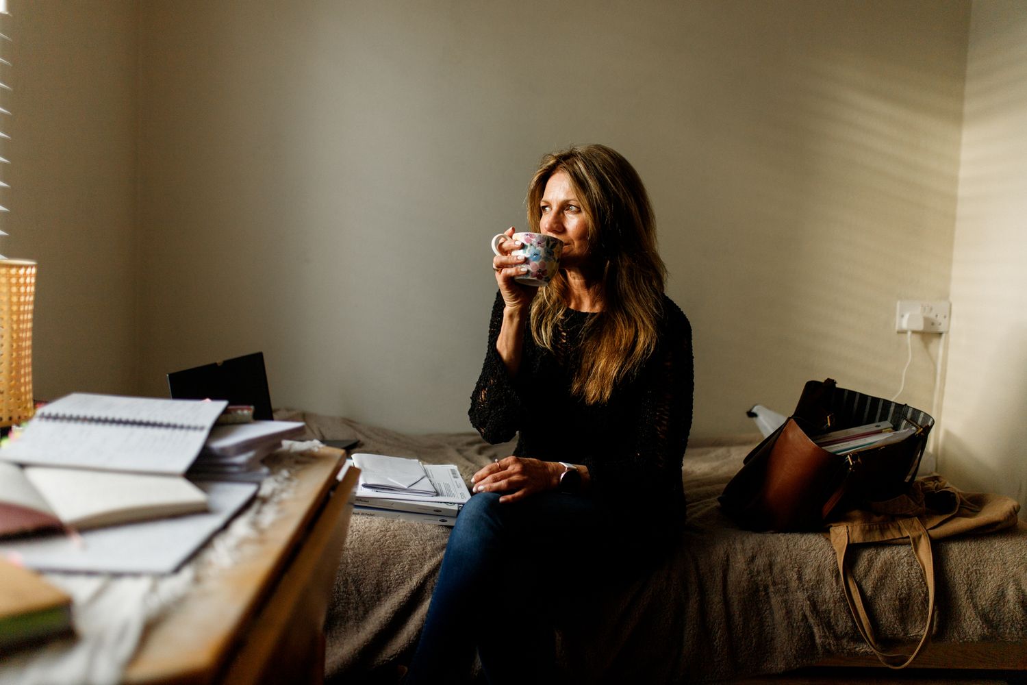 Person in black sitting on bed with papers and books scattered around while drinking from a mug.