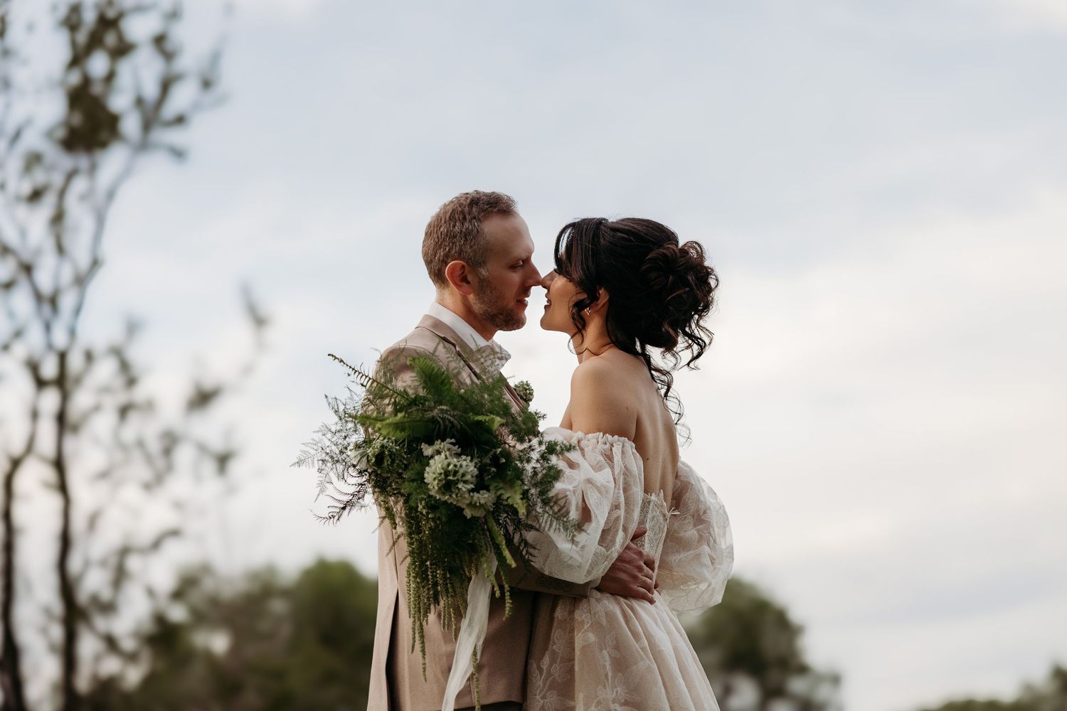 A romantic wedding couple embraces outdoors at sunset while the bride holds a lush greenery bouquet.