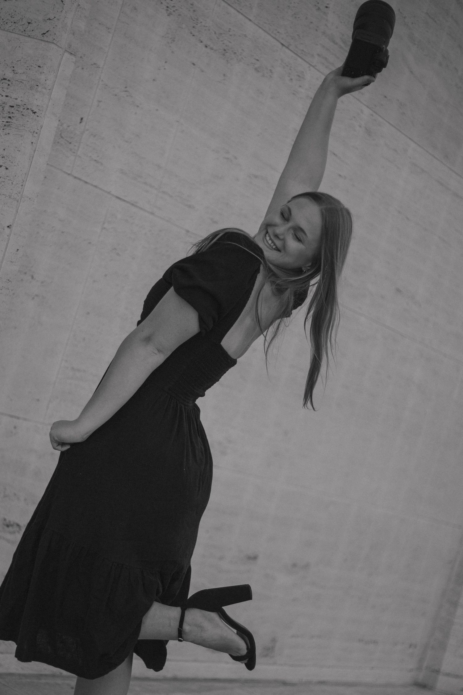 Dancer in a black dress strikes a dramatic tilted pose against a light wall in black and white photography.
