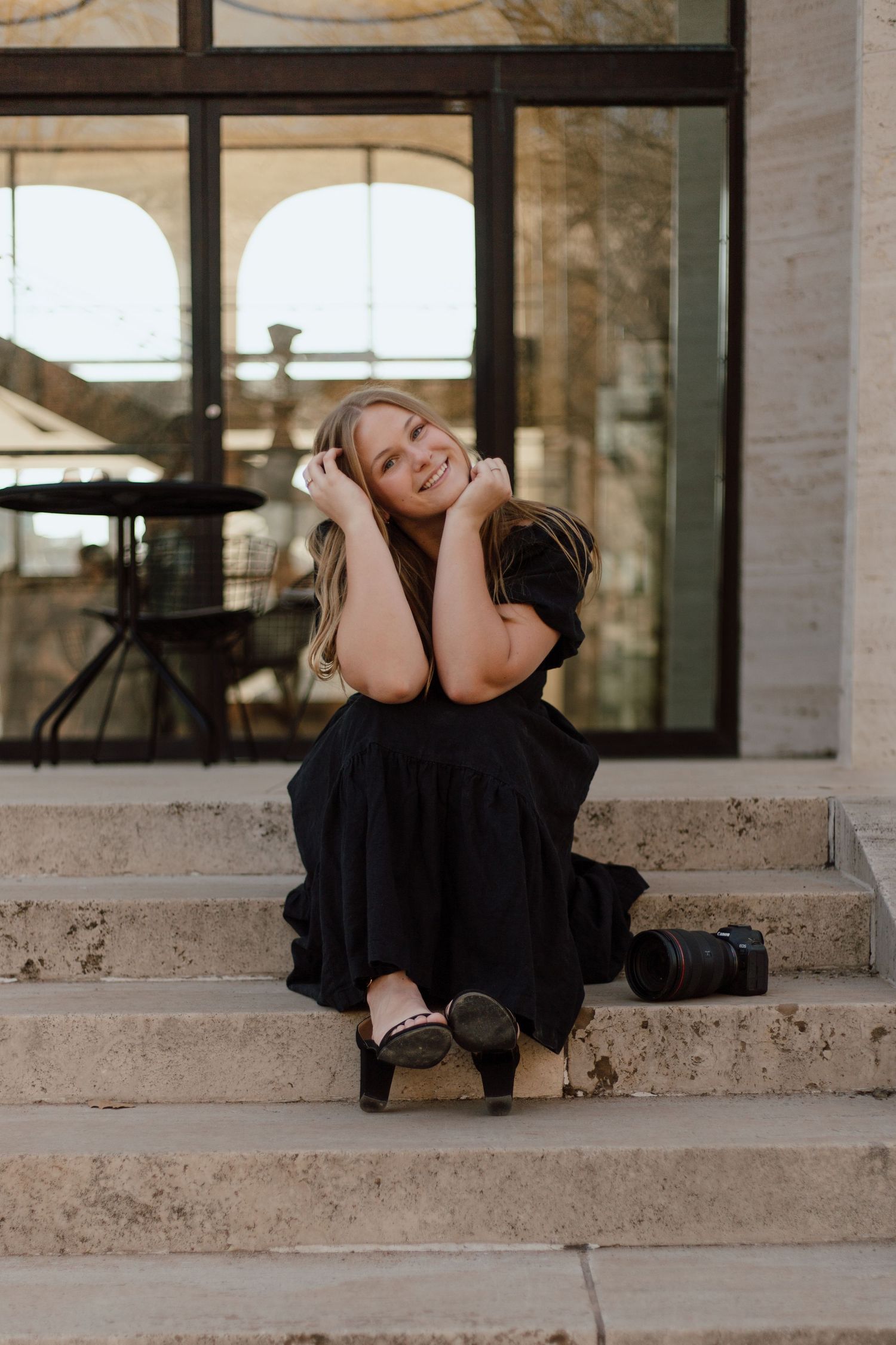 A person in a black dress sits on stone steps outside a building, posing joyfully with hands near their face.
