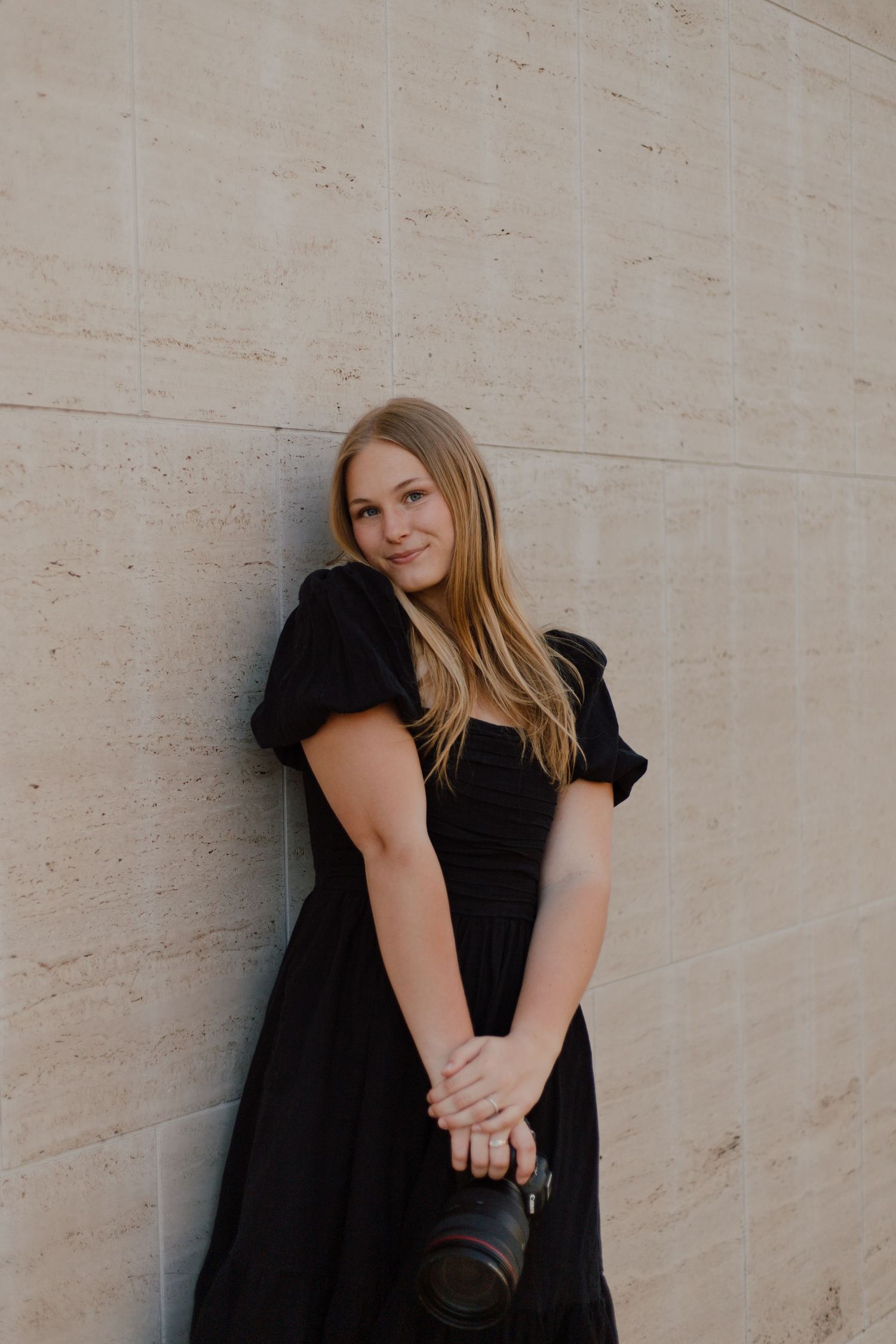 Young woman in black dress holding a camera stands against a beige wall in natural lighting.