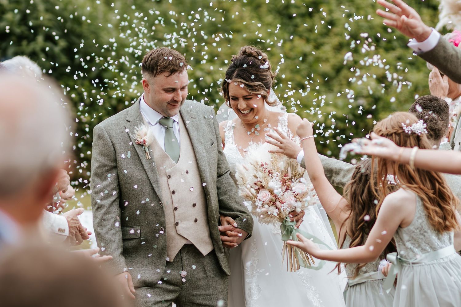 Newly married couple walk through confetti shower surrounded by celebrating wedding guests on sunny day.