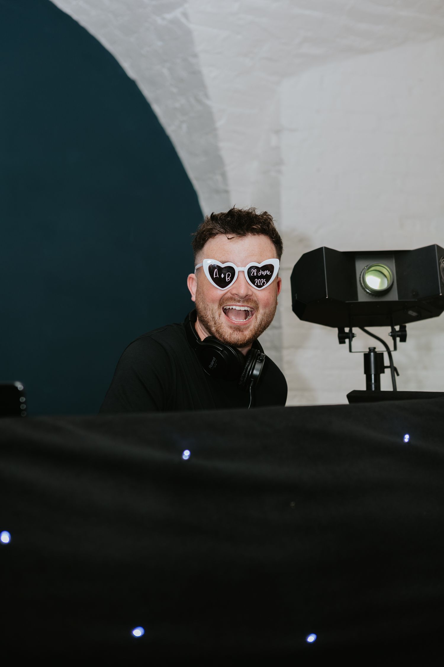 A DJ performs at night behind a deck with heart-shaped sunglasses and equipment visible in silhouette against a dark background.