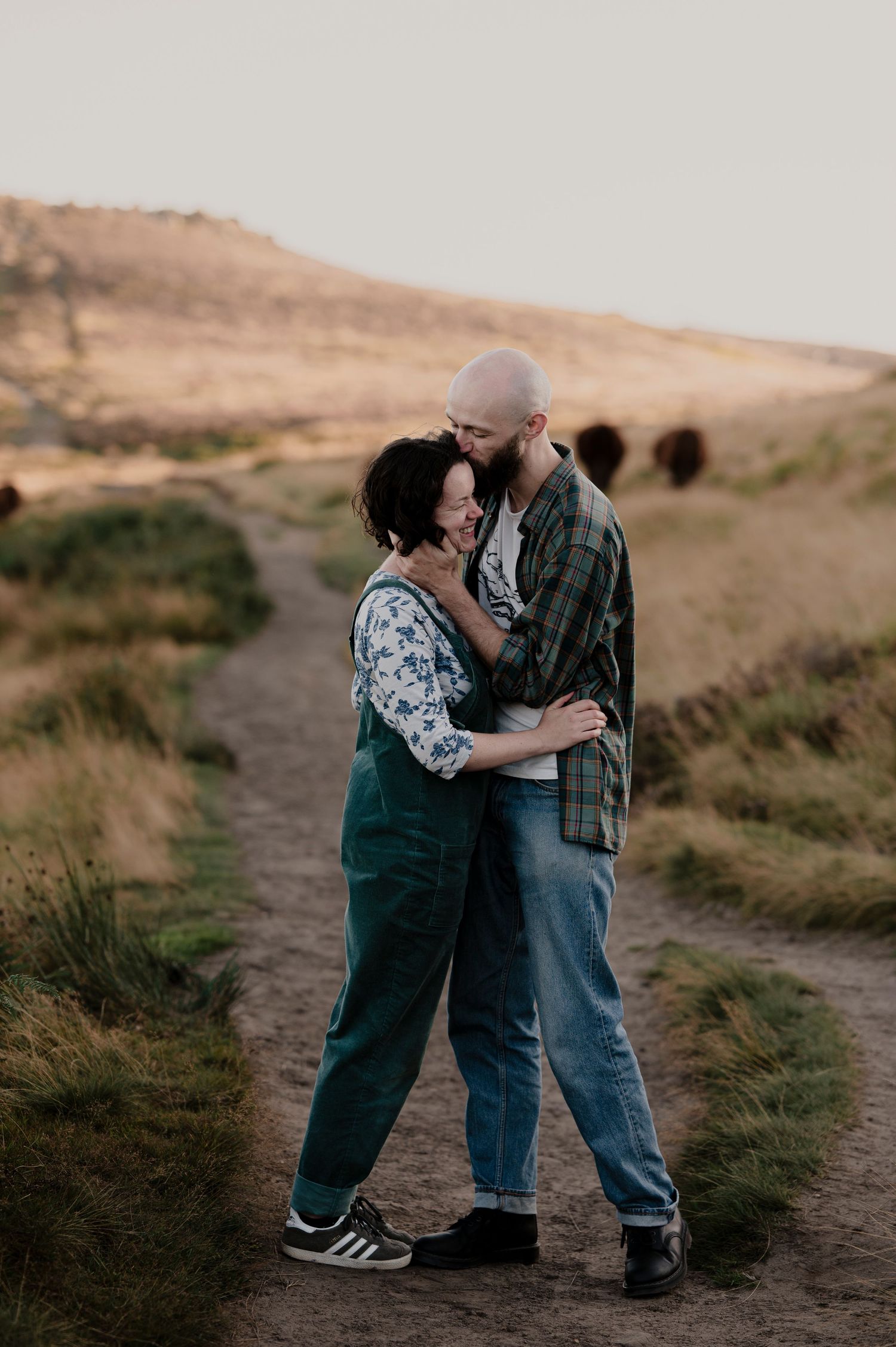 A couple shares an intimate moment on a dirt path surrounded by rolling hills.