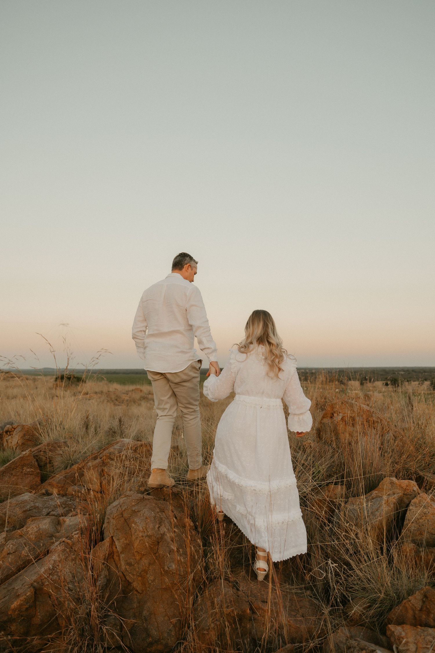 Two people in white outfits hold hands while standing on rocky terrain at sunset against a pale sky.
