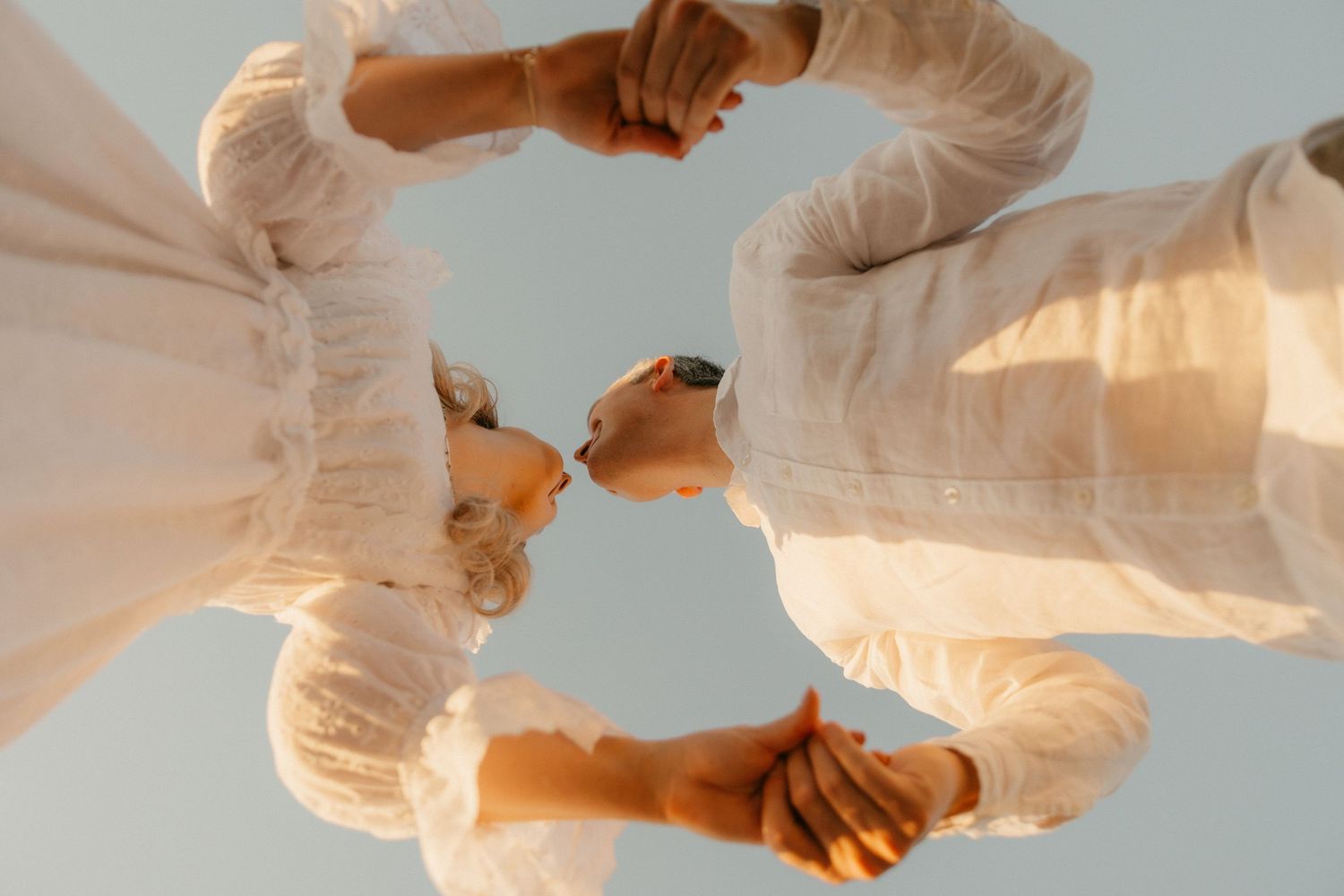 Low angle view of couple in white clothing holding hands in a circular formation against blue sky.
