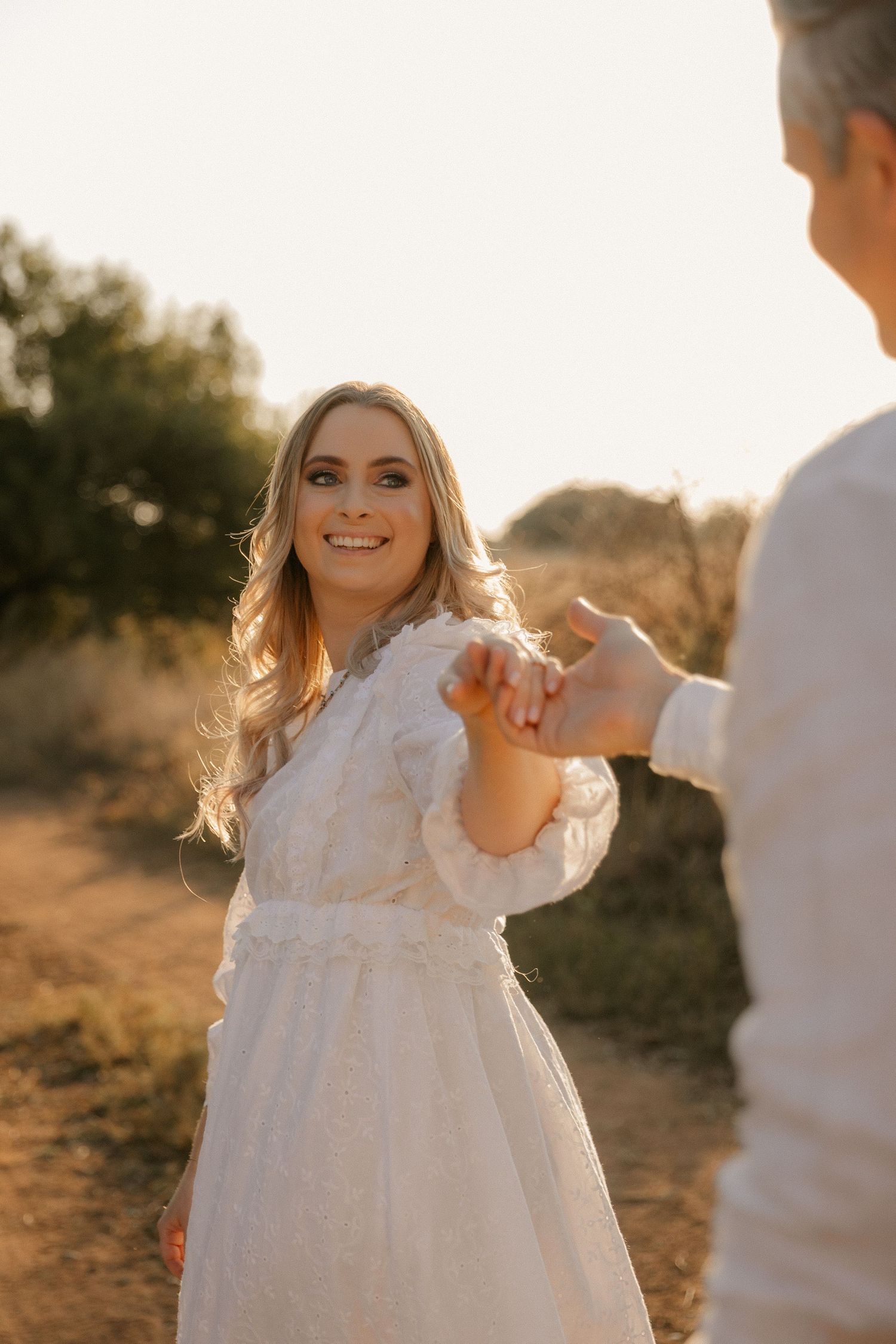 A woman in a flowing white dress smiles while dancing at sunset on a dirt path.
