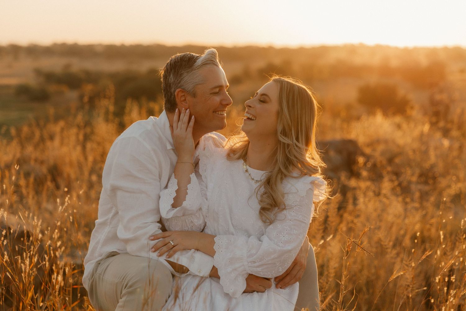 A romantic couple dressed in white embraces in a golden wheat field during sunset, creating a dreamy atmosphere.
