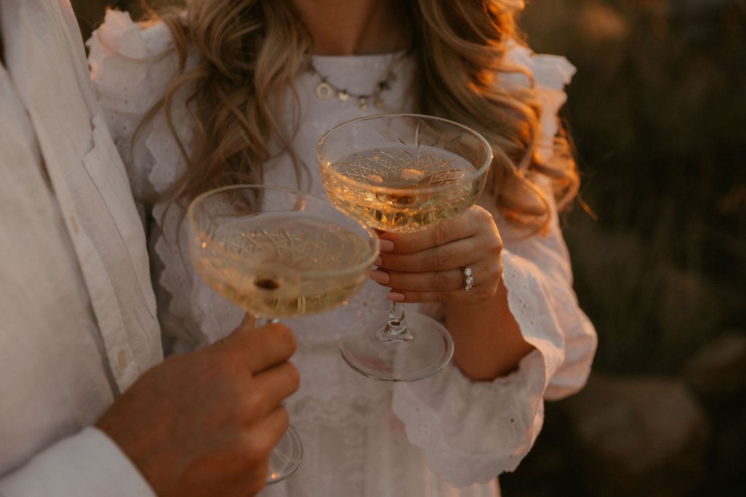 Two hands in white sleeves holding vintage champagne coupe glasses for a celebratory toast at golden hour.