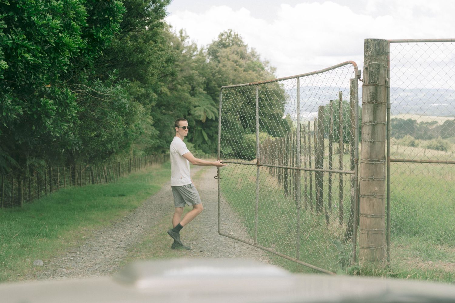 Someone opens a rustic metal gate on a countryside dirt path bordered by green trees and fields.