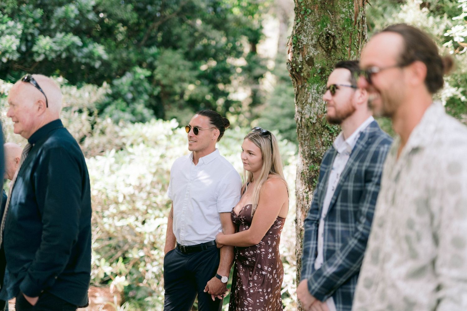 Wedding guests gathered outdoors under trees watching the ceremony.