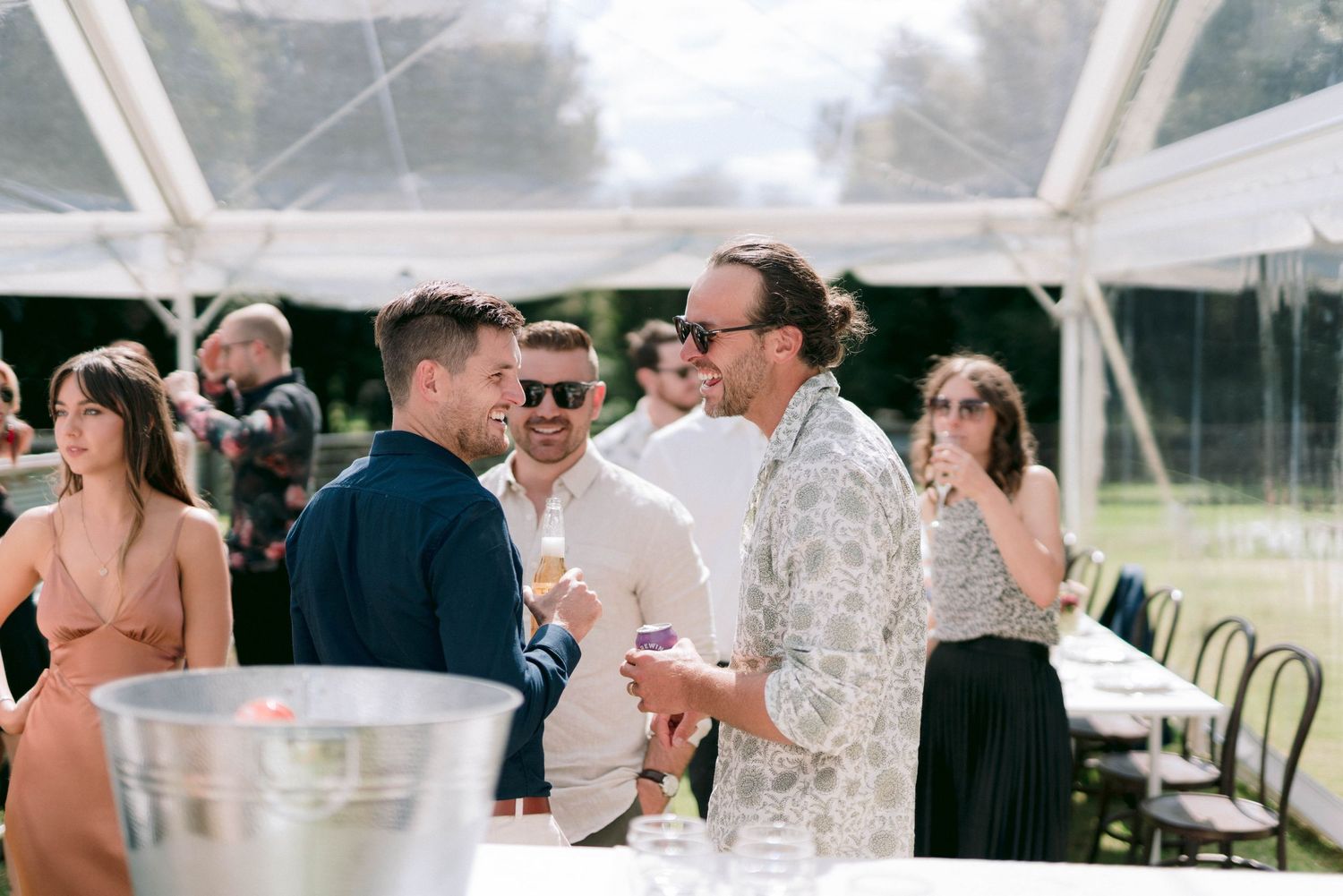 Wedding reception under a white tent with guests mingling at decorated tables.