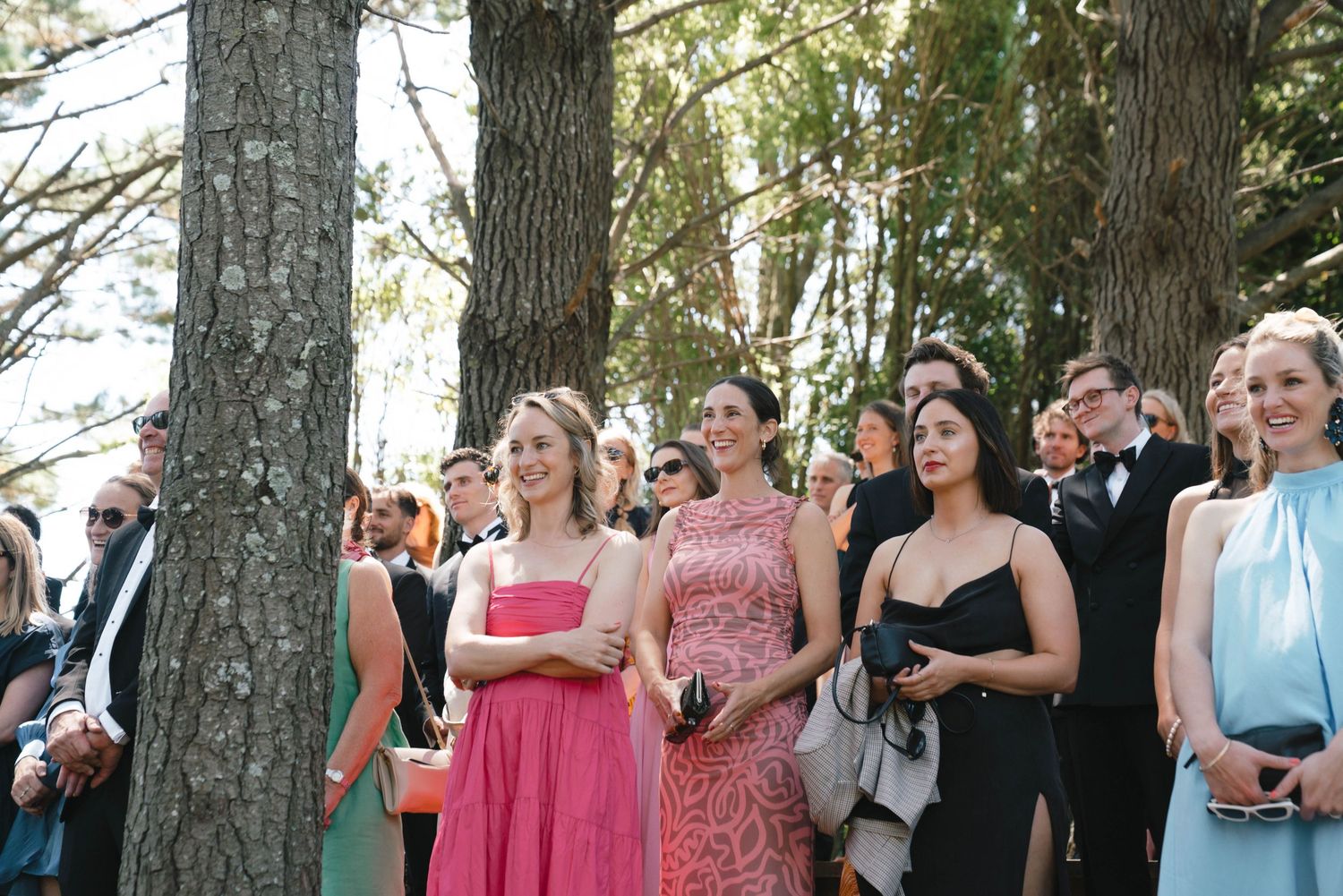 Wedding guests in vibrant dresses observe the ceremony in a wooded outdoor setting.