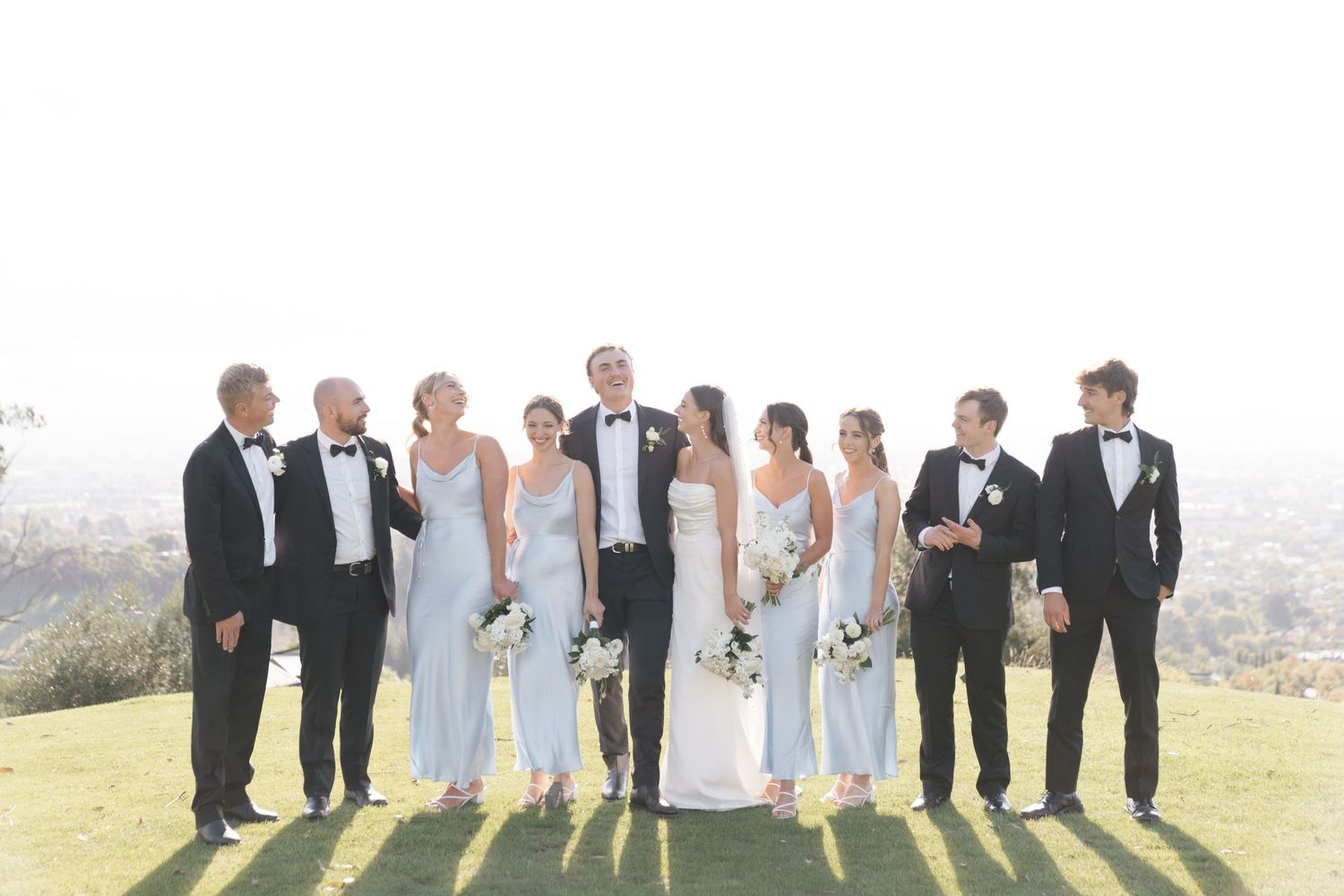 A wedding party stands together outdoors on a hill while posing against a bright sunny background.