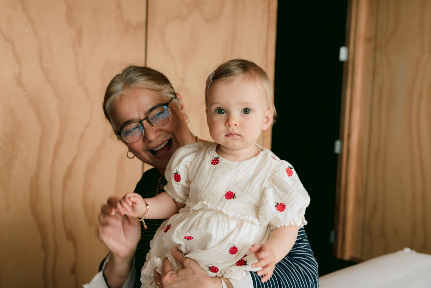Young child wearing a white dress with red heart patterns sits with an adult in front of wooden panel wall.