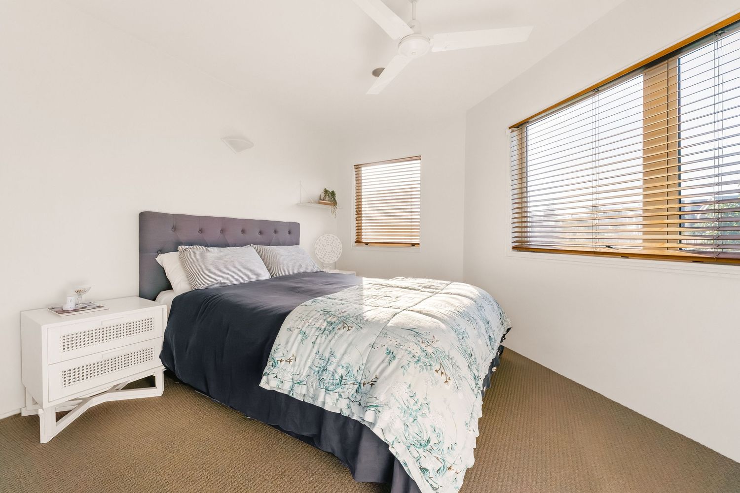 Modern bedroom with grey headboard, white bedside table and floral duvet cover next to window with wooden blinds.
