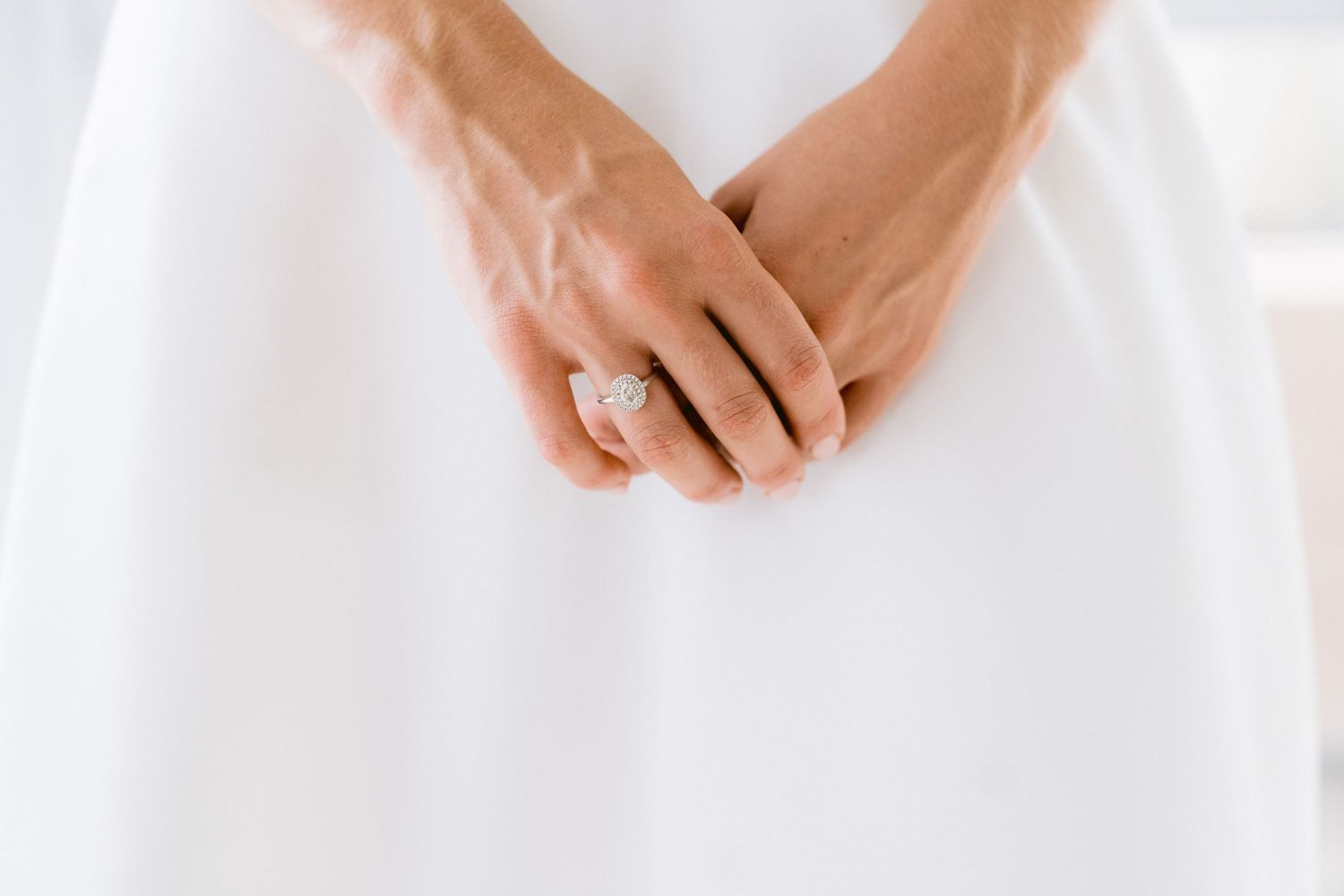 Close-up view of hands wearing a white dress and elegant engagement ring against bright background.