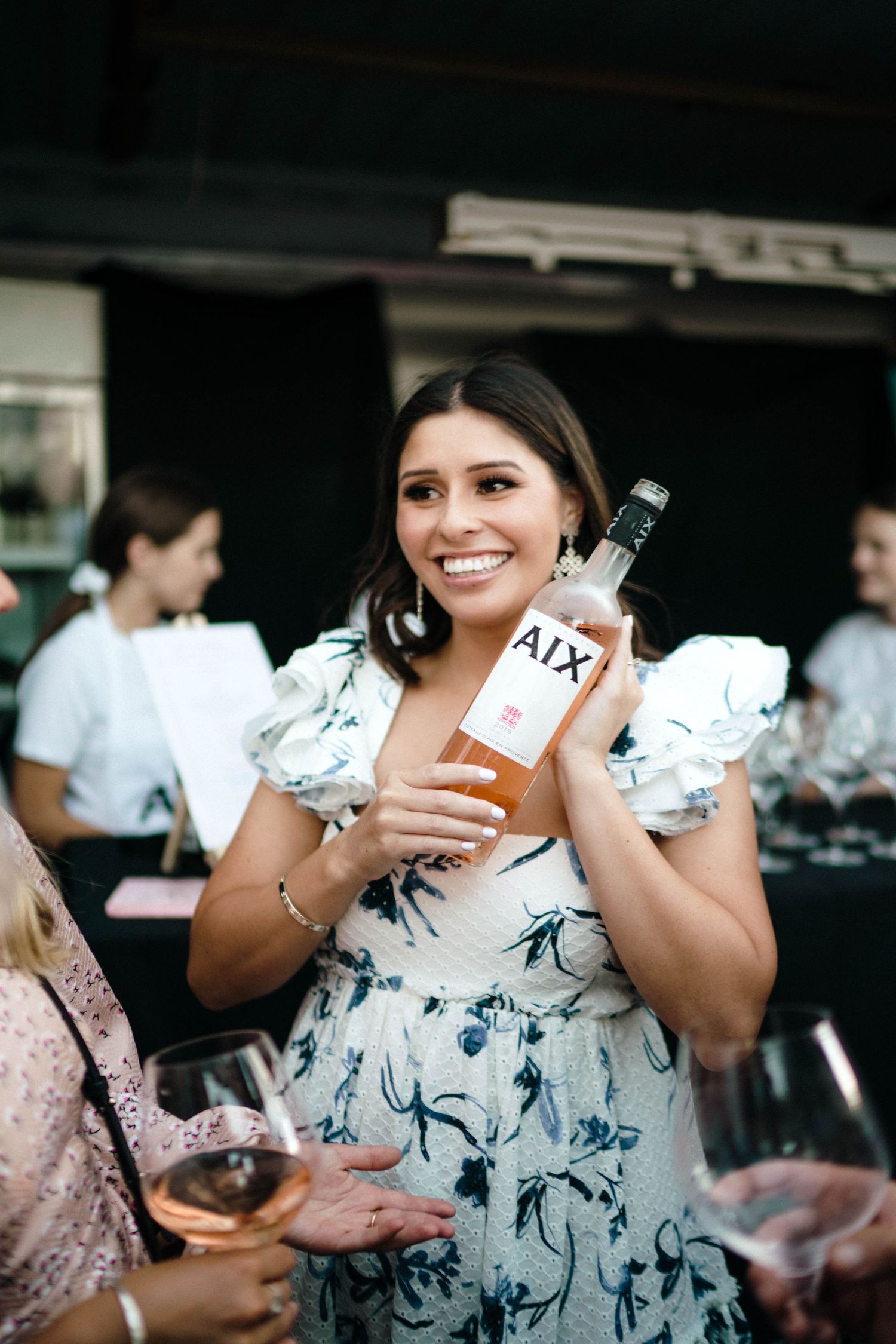 Woman in white floral dress holds a bottle of wine at a social gathering outdoors.