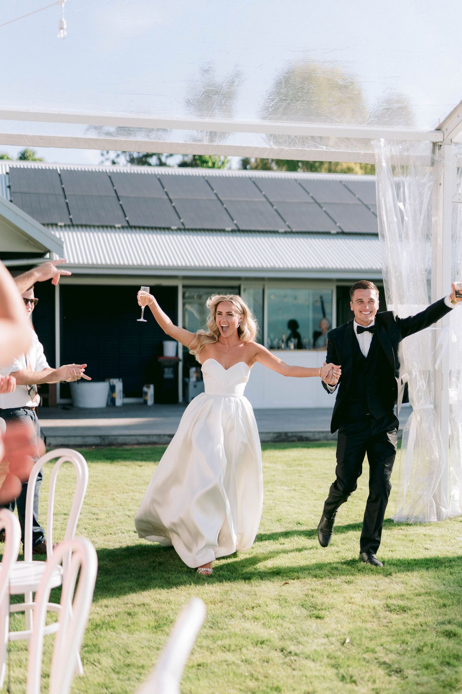 A joyful wedding celebration on a lawn as the newlyweds make their grand entrance under a white tent.