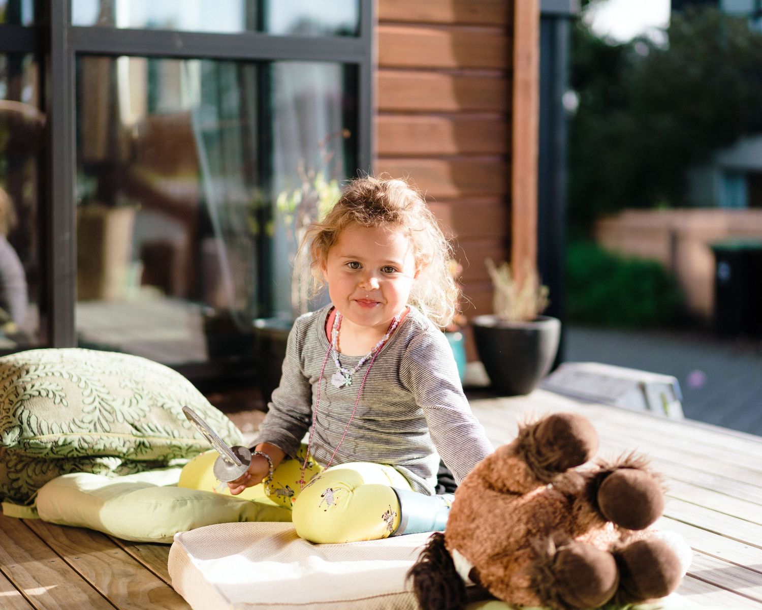 A toddler sits on a patio with pillows and a stuffed animal in casual daytime lighting.