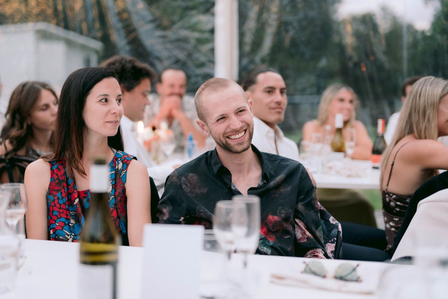 Wedding reception guests enjoy dinner and conversation under festive lighting at an outdoor table setting.