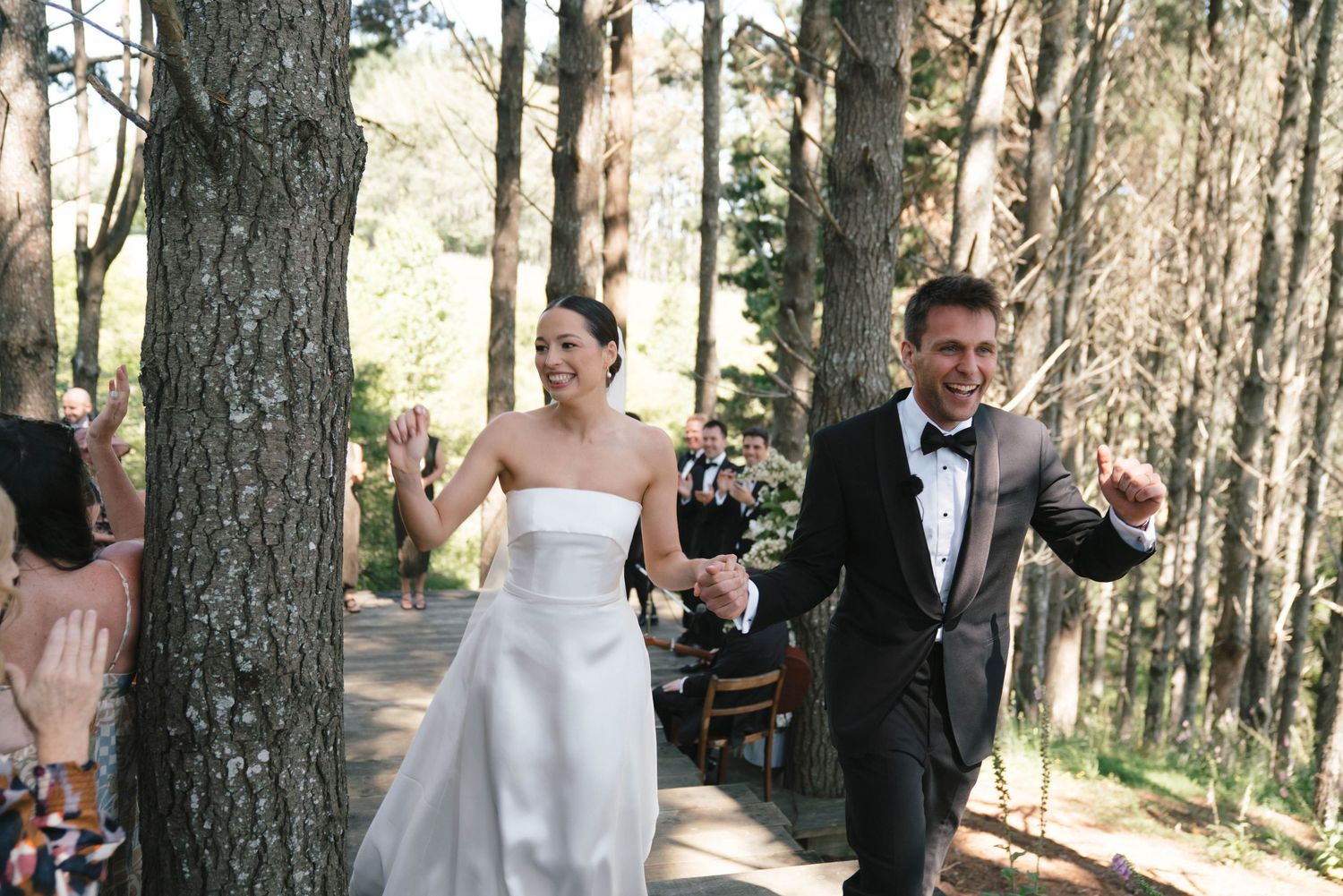 A wedding party gathers for photos in a sunlit wooded area with tall pine trees creating a natural outdoor setting.