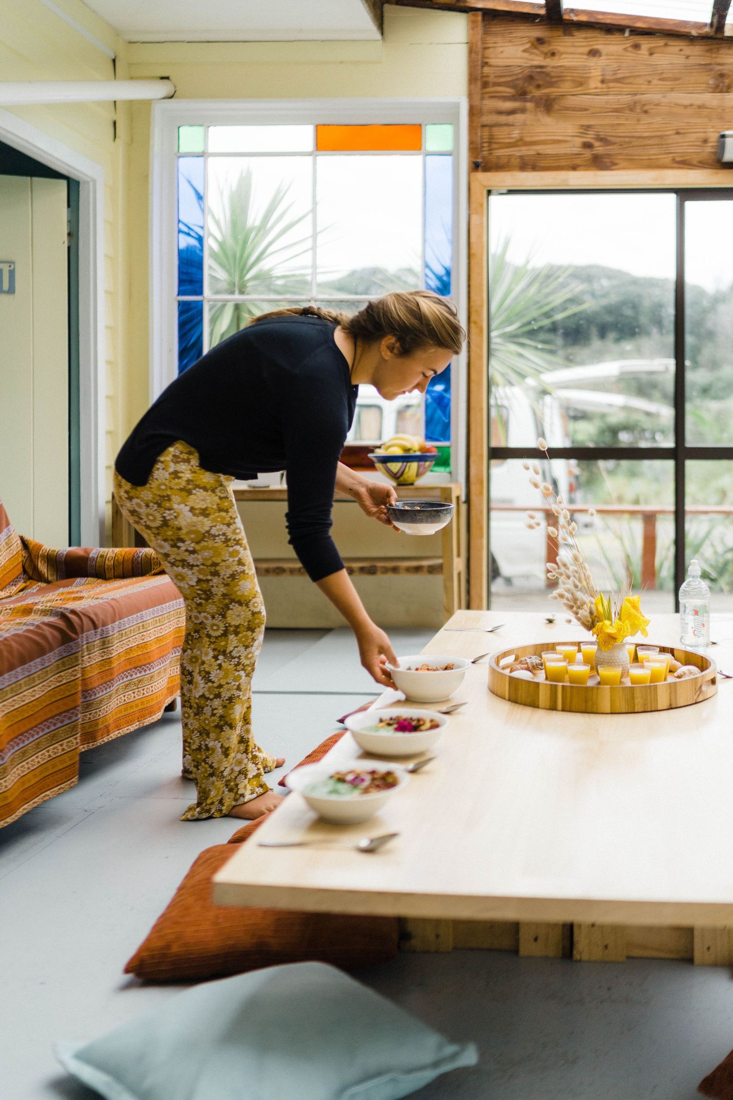Person arranging food items on a long wooden table in a modern room with large windows.