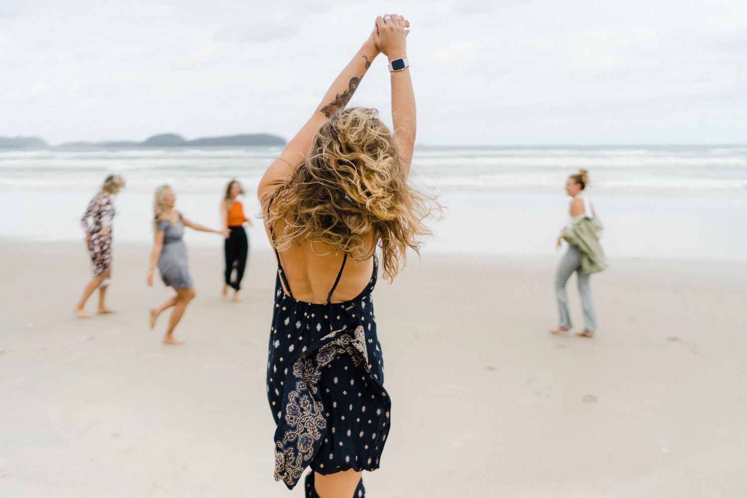 Person dancing on beach with arms raised while others walk in background.