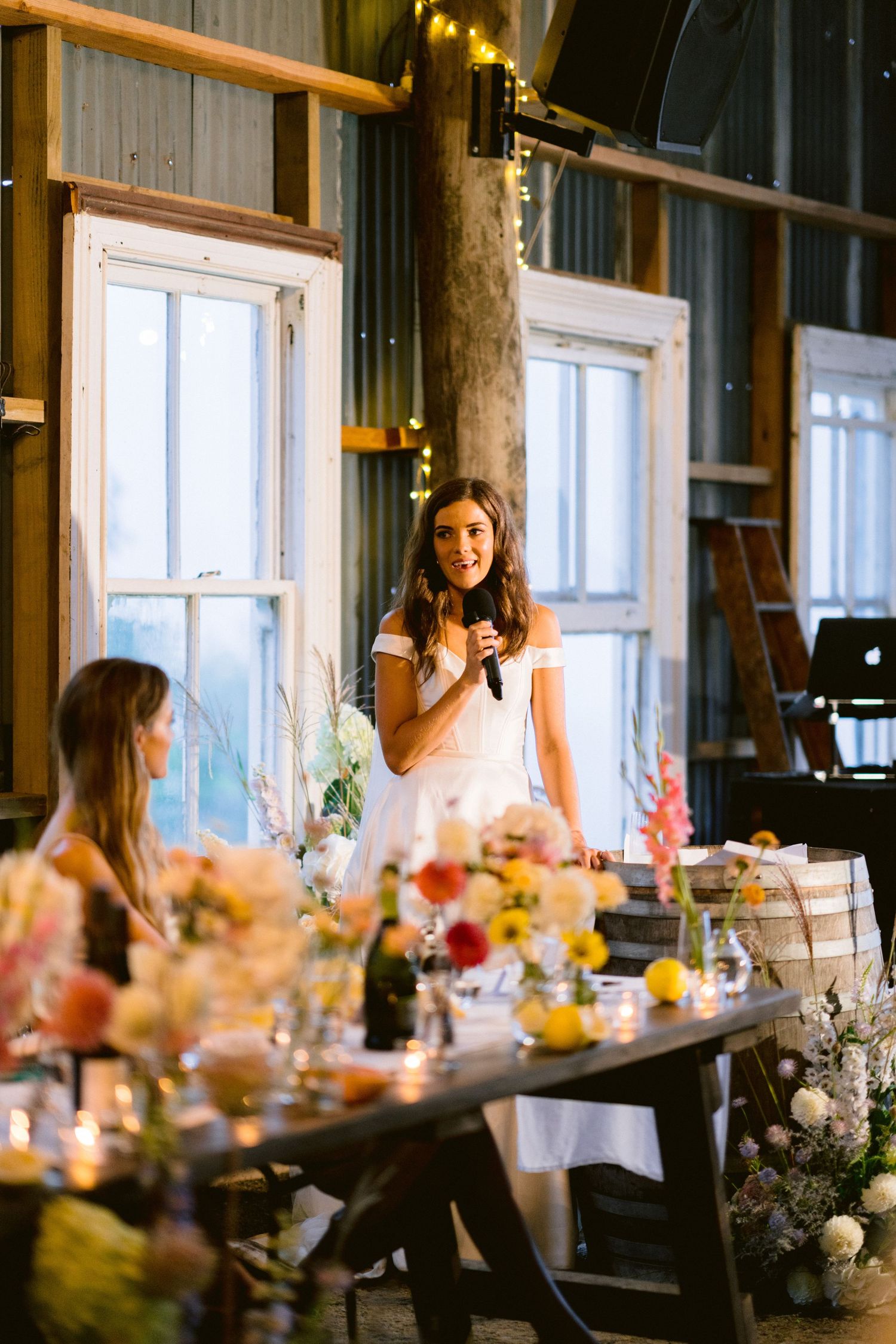 A wedding reception speech being given in a rustic venue with string lights and floral decorations.