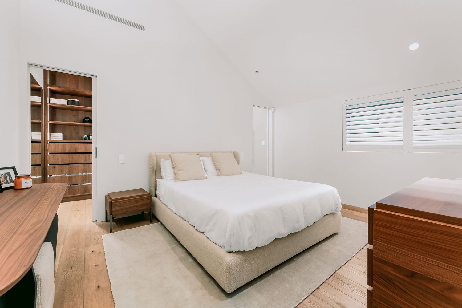 Modern minimalist bedroom with beige platform bed, wooden desk, and built-in shelving in bright white interior.